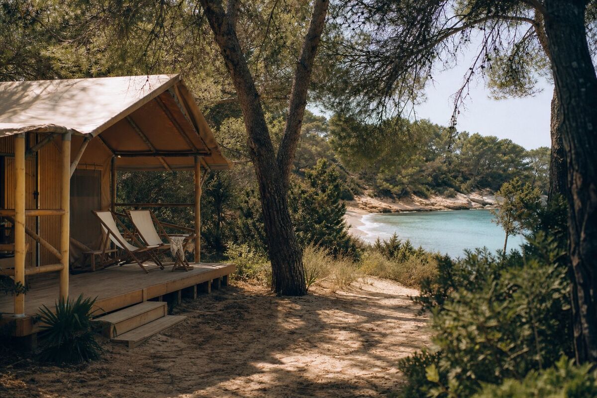 Wooden camping bungalow with canvas roof among pines, sandy path leading to a turquoise beach on Ibiza's east coast