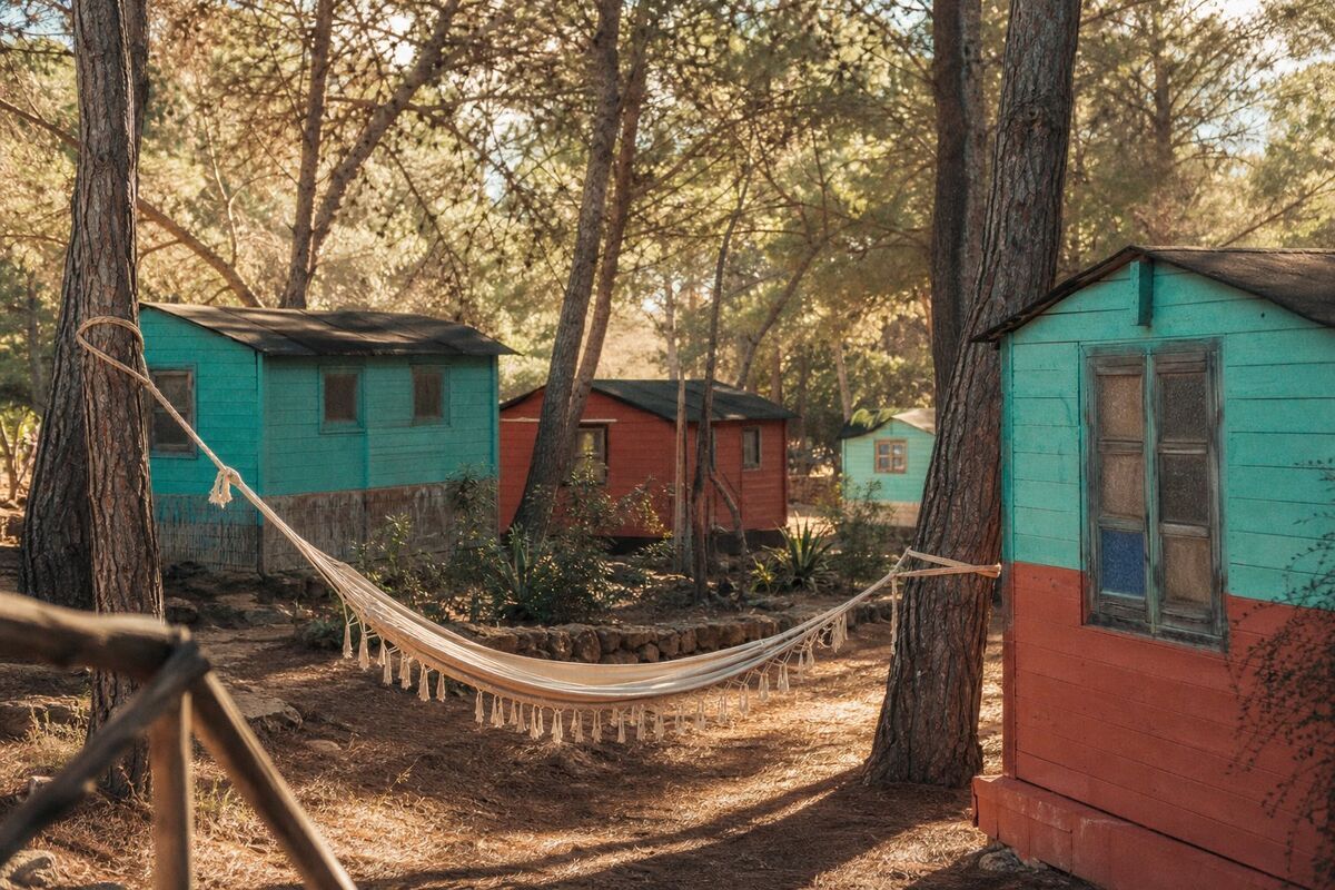 Colourful small wooden camping cabins in faded turquoise and terracotta scattered among tall pine trees, with a hammock between the pines